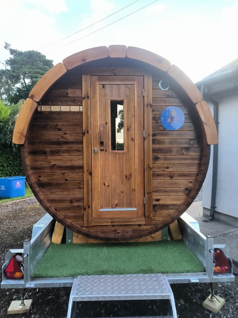 A wooden barrel-shaped sauna on a trailer sits outdoors next to a building.