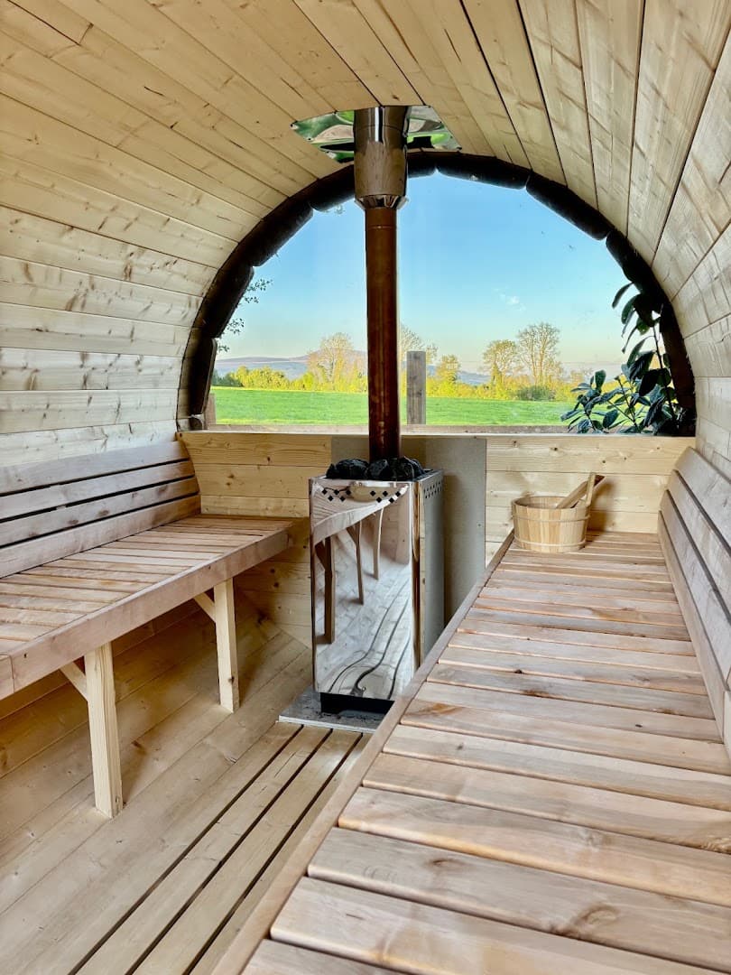 Interior view of a wooden barrel sauna with a metal stove, benches, and a large window overlooking a grassy landscape.