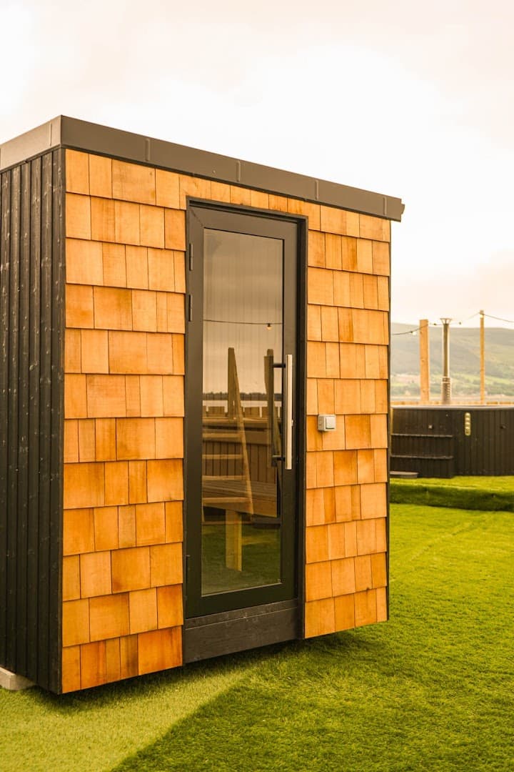A wooden sauna structure stands on a grassy lawn near the sea.
