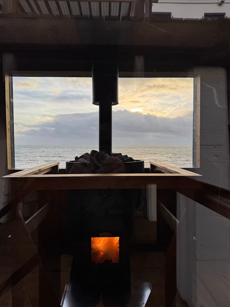 A wood-burning stove sits inside a seaside sauna, framing an ocean view through a window.