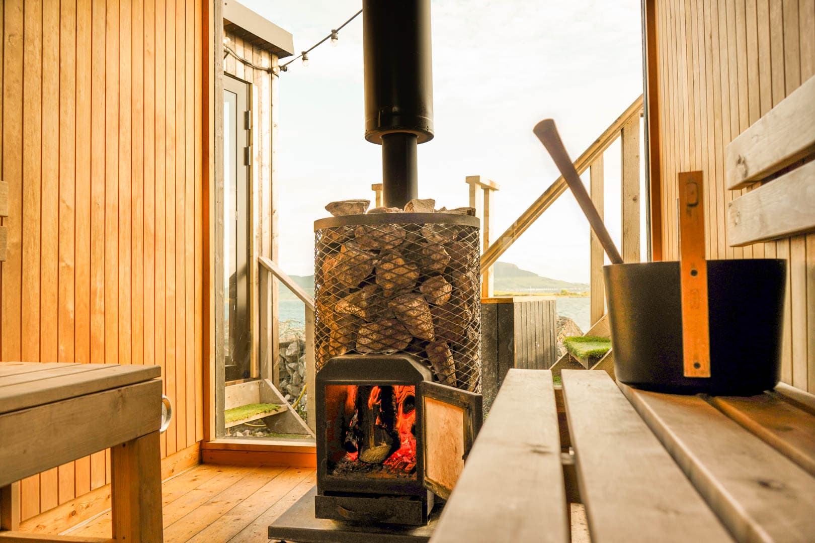 A wooden sauna interior frames a lit wood-burning stove and a bucket, with an ocean view outside.