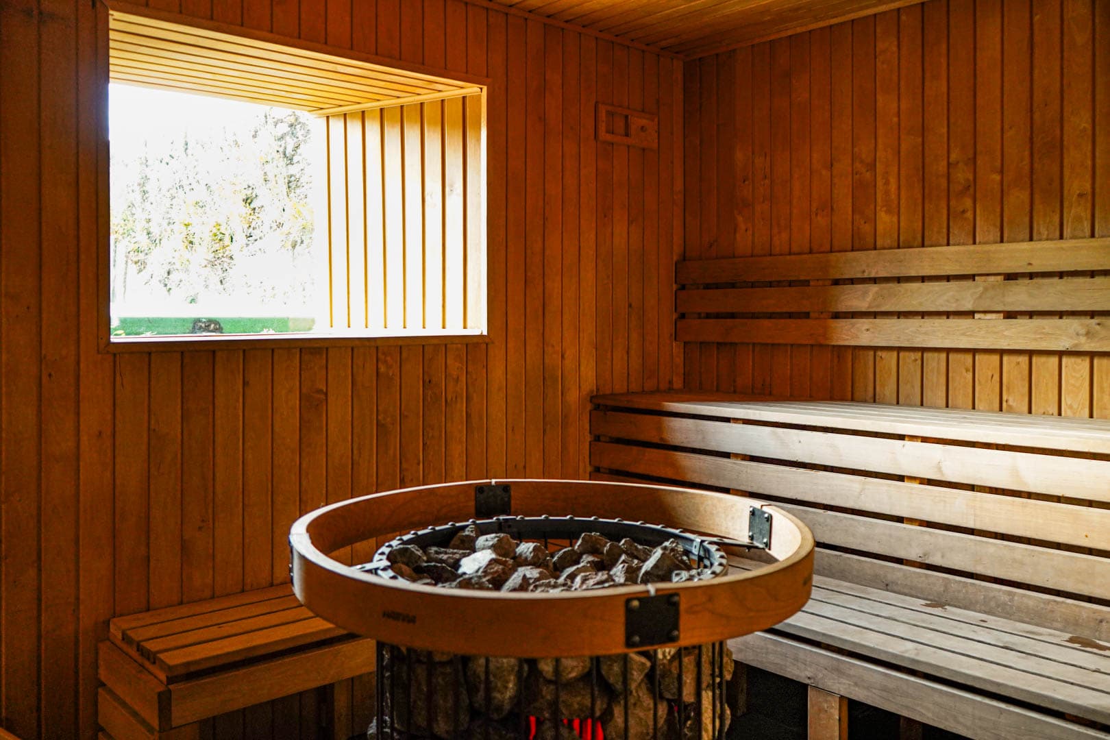 Interior view of a wooden sauna featuring a stone stove and tiered bench seating next to a window.