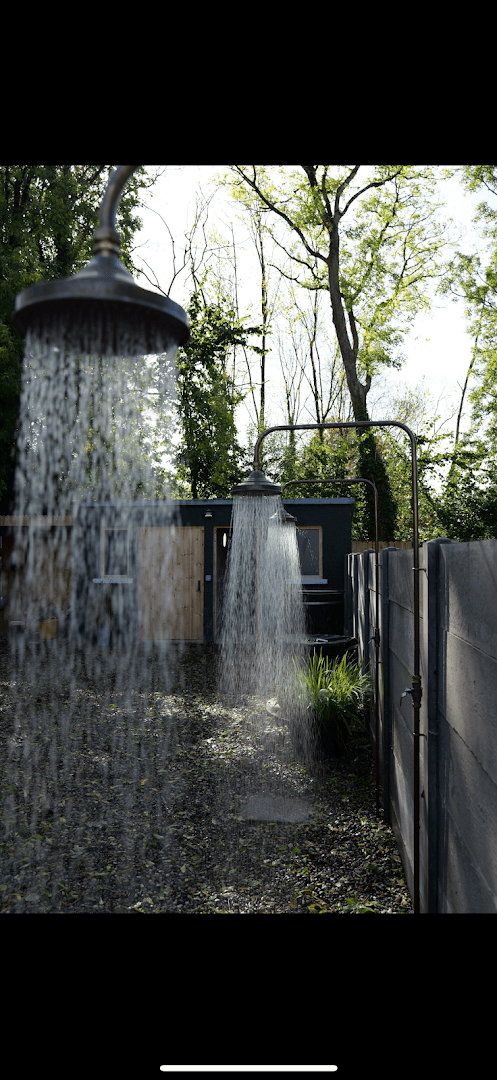Three outdoor shower heads spray water in a rustic backyard setting with a wooden shed and trees.