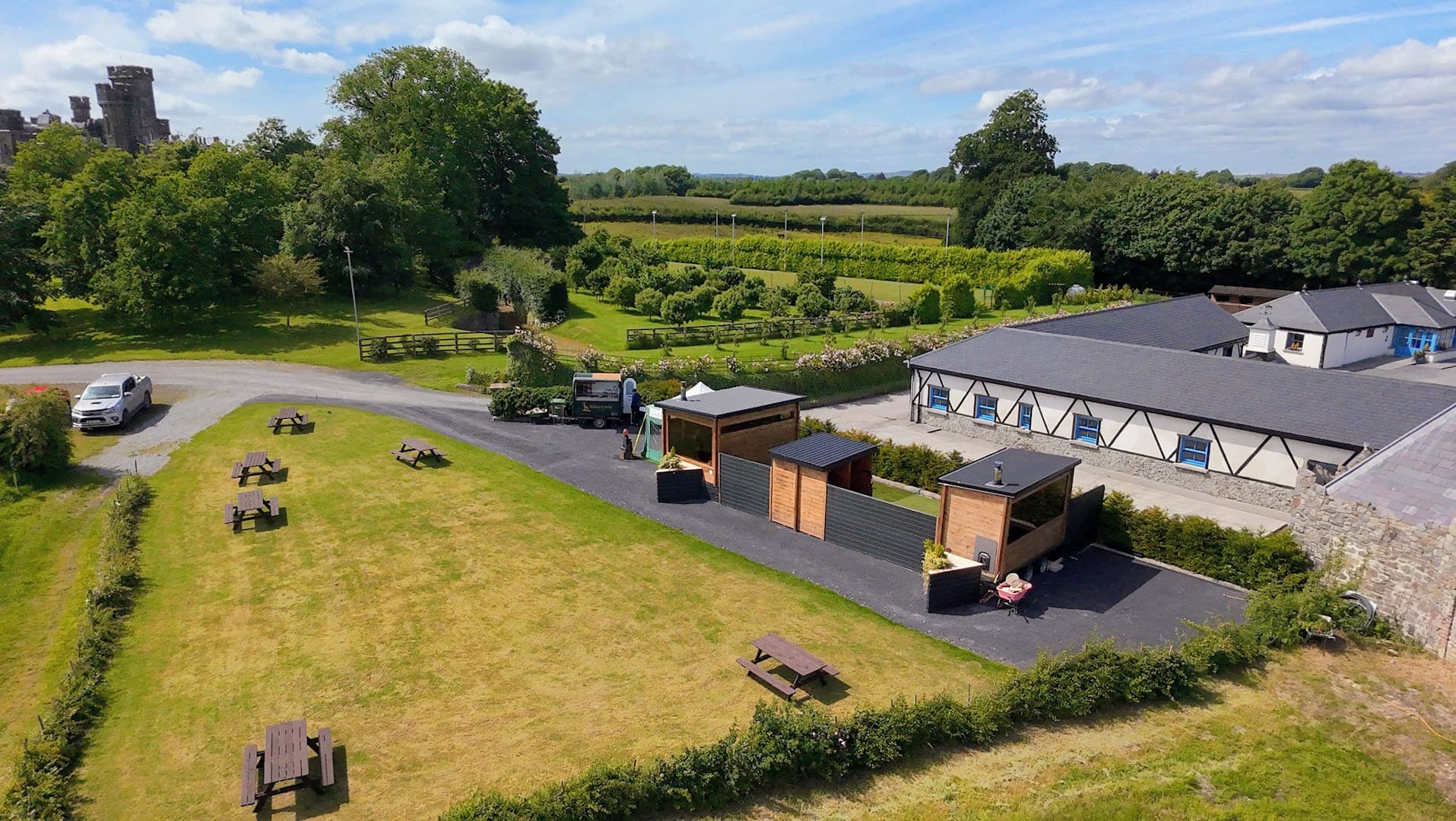 An aerial view shows three wooden sauna structures on a paved path next to a grass lawn and buildings.
