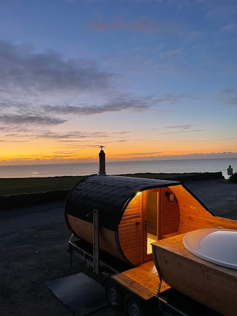 A barrel sauna with a hot tub on a trailer sits by the sea at sunset.