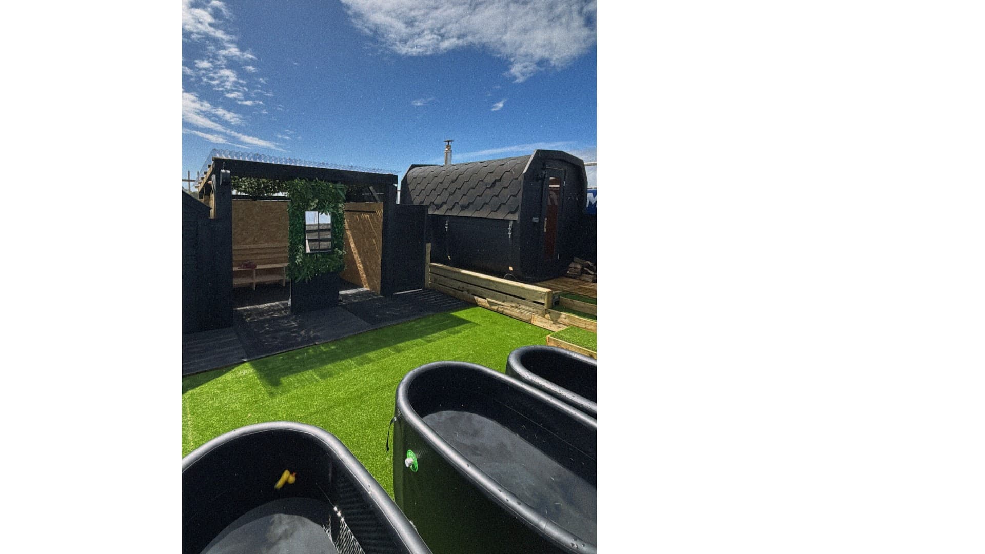 Black sauna cabin and cold plunge tubs outdoors on artificial grass under a sunny blue sky.