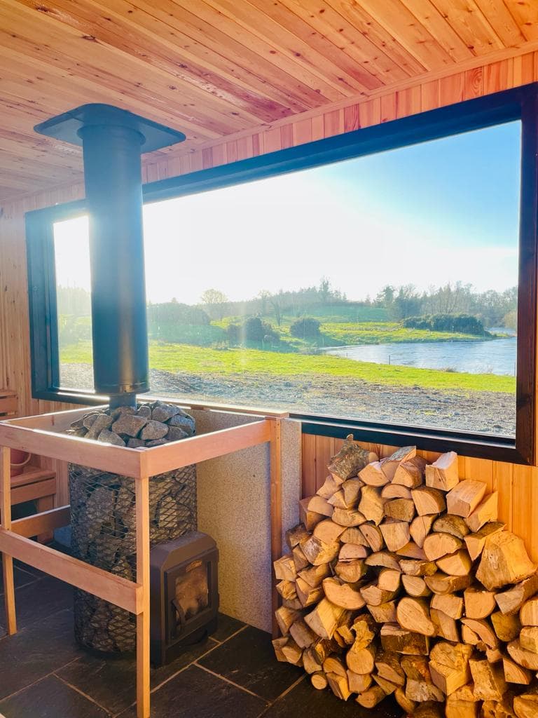 Inside a wooden sauna with a large picture window, a stone heater with a chimney stands beside a stack of firewood.