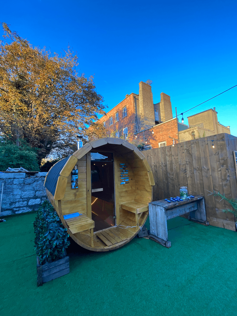 Wooden barrel sauna with a glass door sits on artificial grass in an outdoor area next to a brick building.