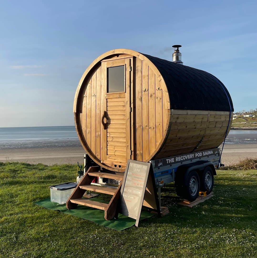 A wooden barrel sauna on a trailer sits by a grassy beach overlooking the sea.