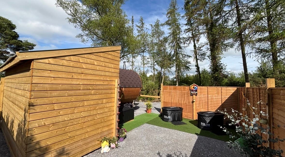 Wooden sauna structure and cold plunges stand in an outdoor enclosure surrounded by gravel, turf, and trees.