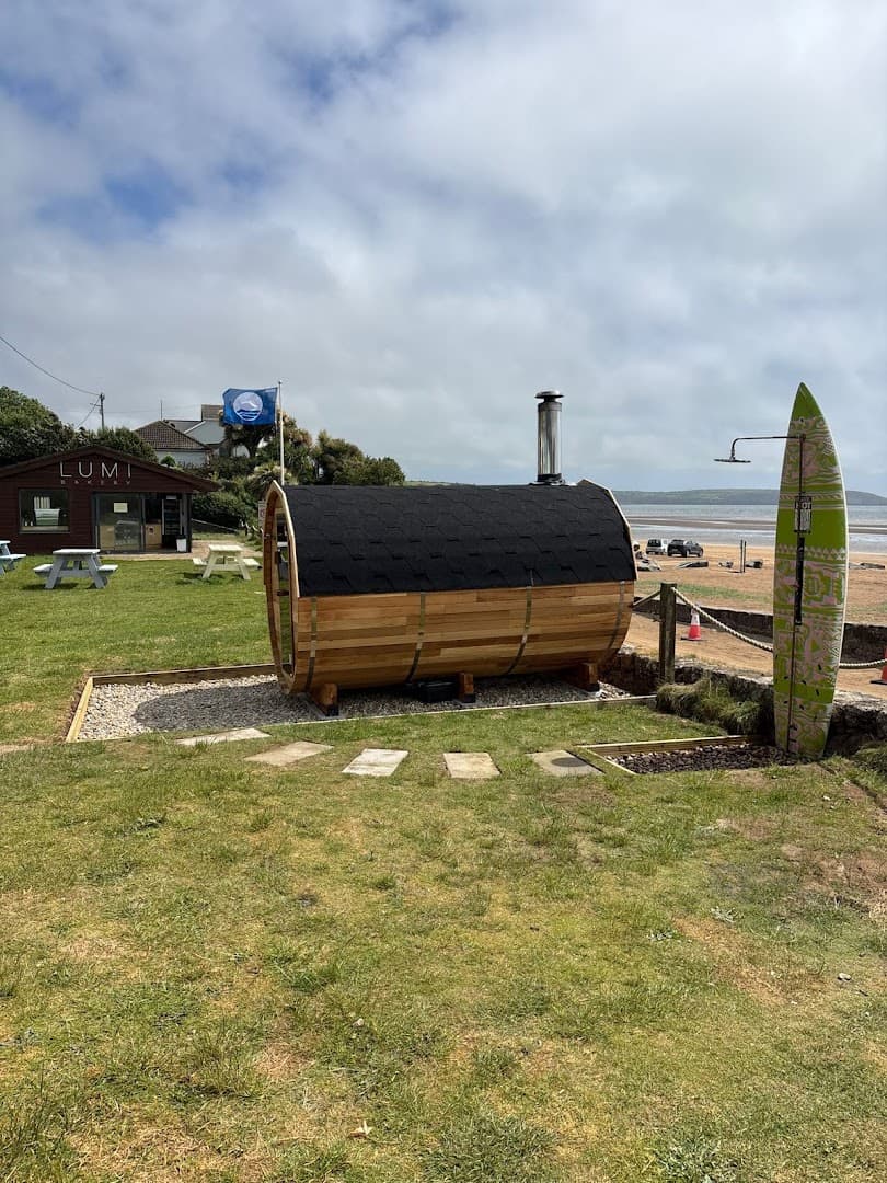 A barrel-shaped wooden sauna with a black shingle roof sits on a grassy area near the sea.