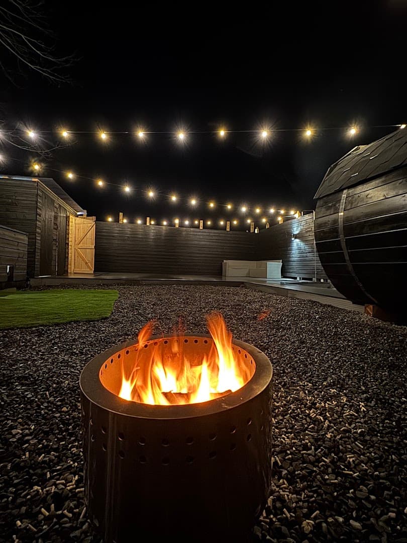 Nighttime view of a fire pit, a wooden barrel sauna, string lights, and an outdoor seating area.
