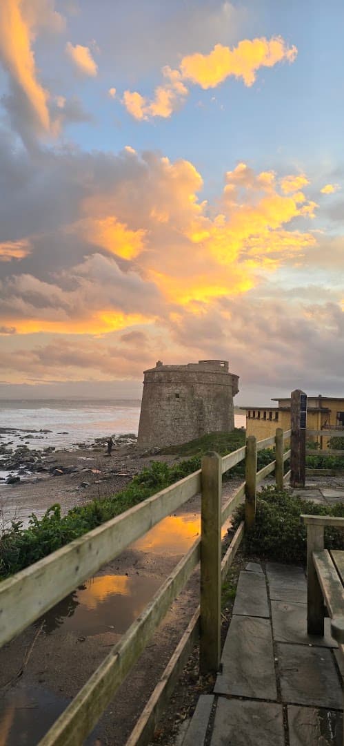 Sunset sky with orange clouds above a coastal tower, a wooden fence, and a stone path near the sea in Donabate.