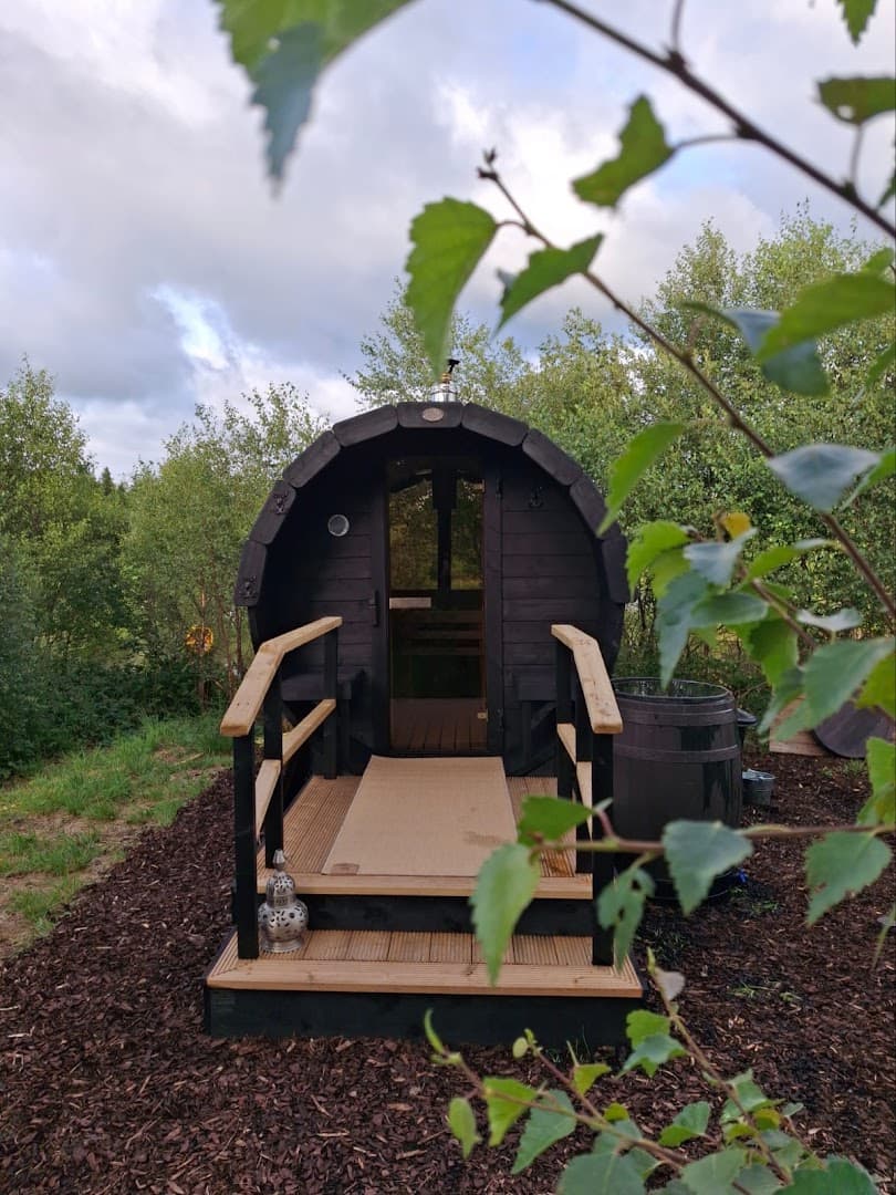 An outdoor barrel sauna stands amidst trees under a cloudy sky.
