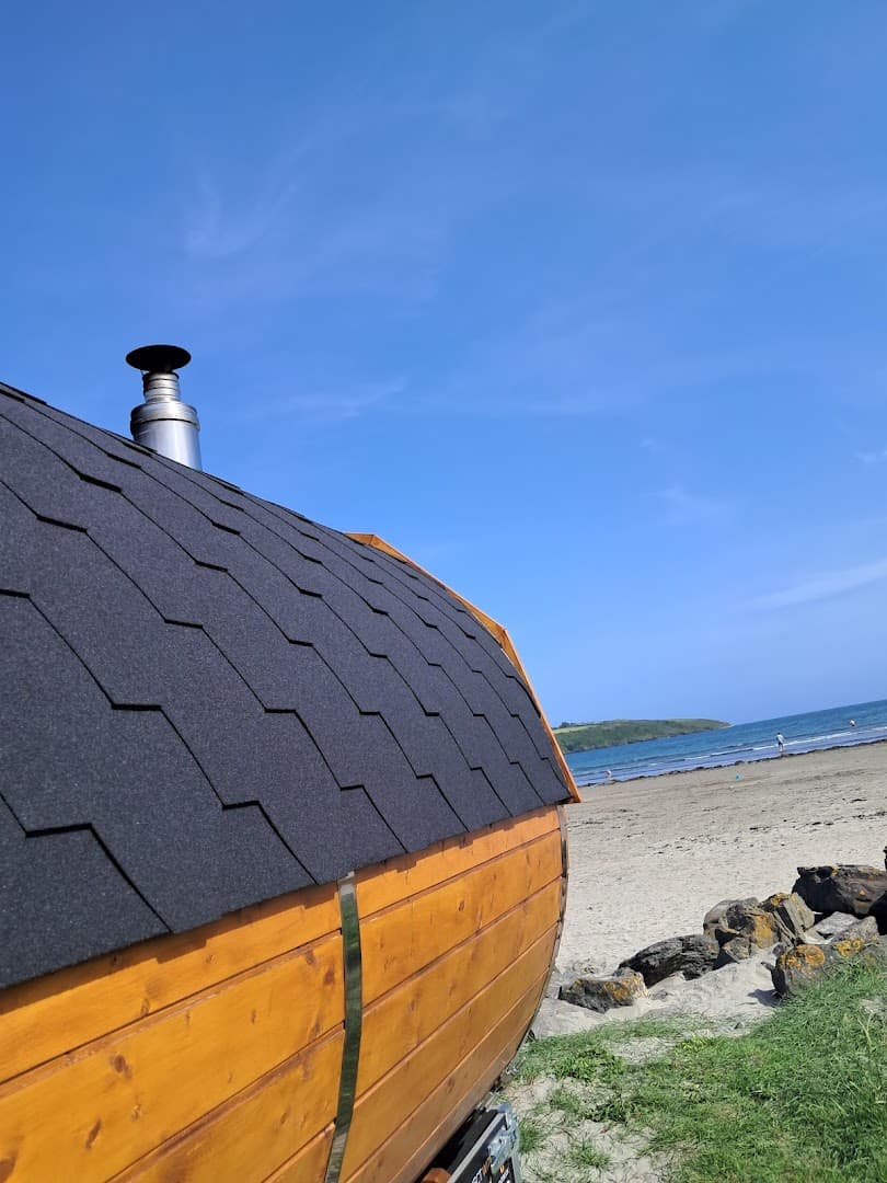 A wooden barrel sauna on a sandy beach with a metal chimney and the sea in the background.