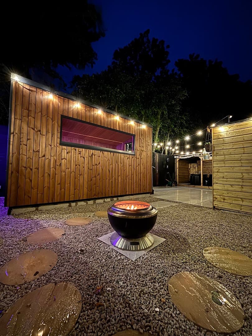 Outdoor wooden sauna building at dusk with a lit fire pit on gravel ground.