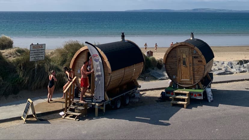 Two barrel-shaped mobile saunas sit near a sandy beach and blue sea, with people standing outside near wooden steps.