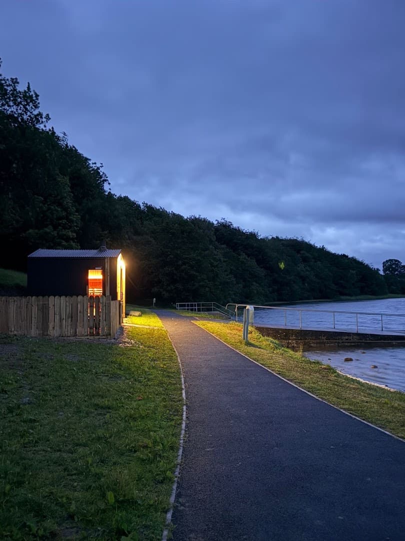 A small dark sauna building glows on a paved path next to a body of water at dusk.
