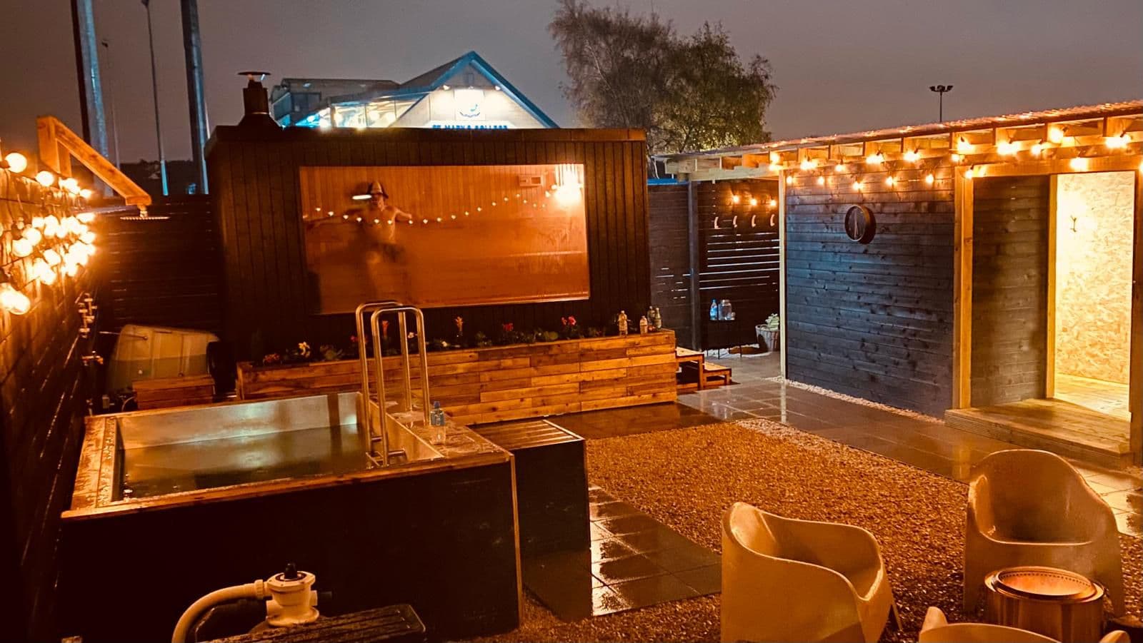 At night, two light-strung dark wooden buildings enclose a gravel courtyard with a cold-plunge tub and a glowing sauna window showing a silhouette inside.