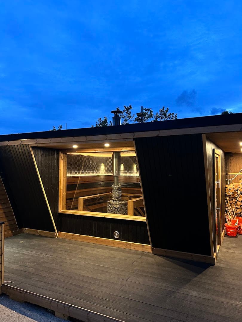 Exterior view of The Sauna Village at dusk, featuring a modern wooden sauna structure with a large glass window and a stone stove.