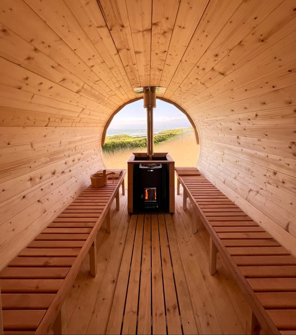 Inside a wooden barrel sauna, two slatted benches flank a central wood-burning stove facing a large window overlooking the sea.