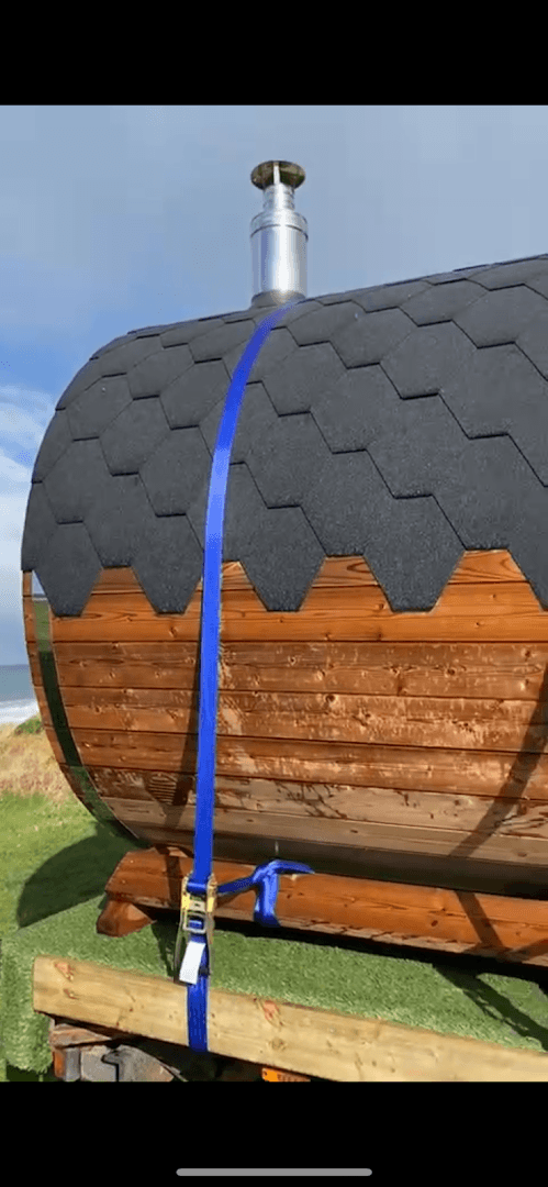 A barrel sauna with a chimney is secured with a blue strap on a trailer near the sea.