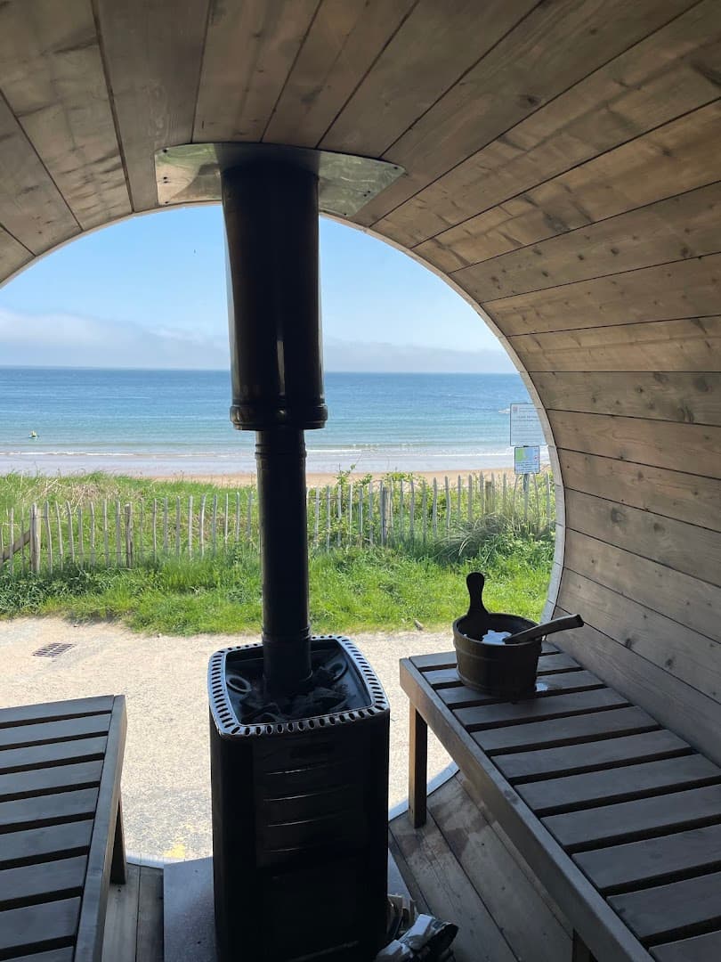 Inside a barrel sauna, a wood stove faces a large arched window with a view of the sea and beach.