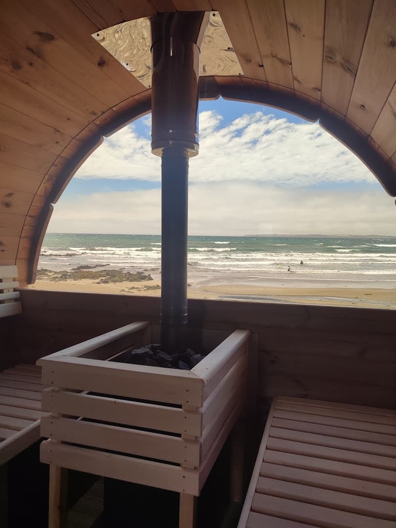 Interior view from a wooden sauna looking out through a large window toward the ocean and beach, with people swimming in the distance.