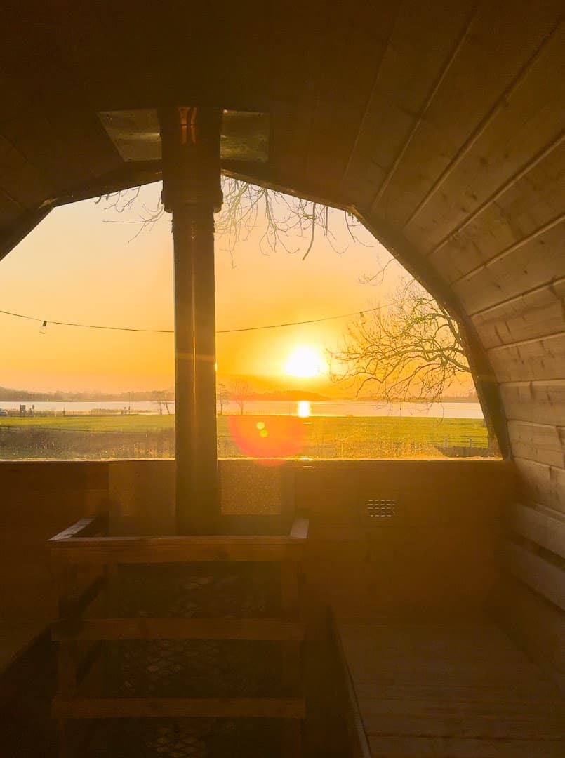 Inside a wooden sauna with a large window overlooking a body of water and a sunset.