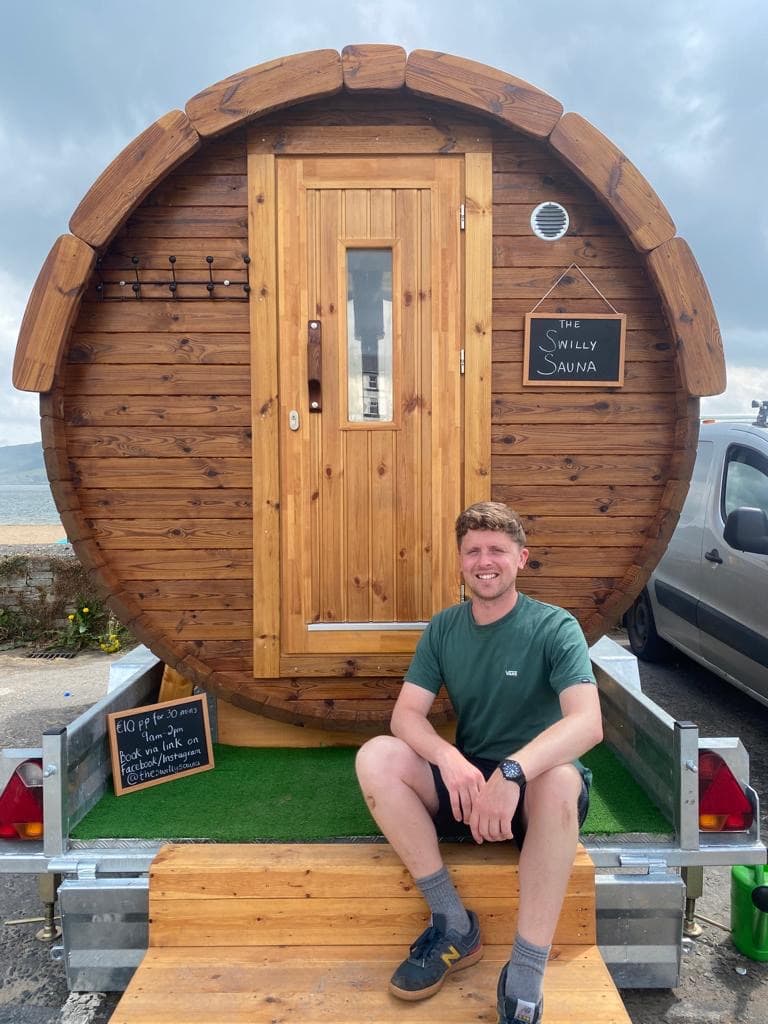 A young man wearing a green T-shirt sits on the wooden steps of a barrel-shaped mobile sauna named "The Swilly Sauna" by the sea.