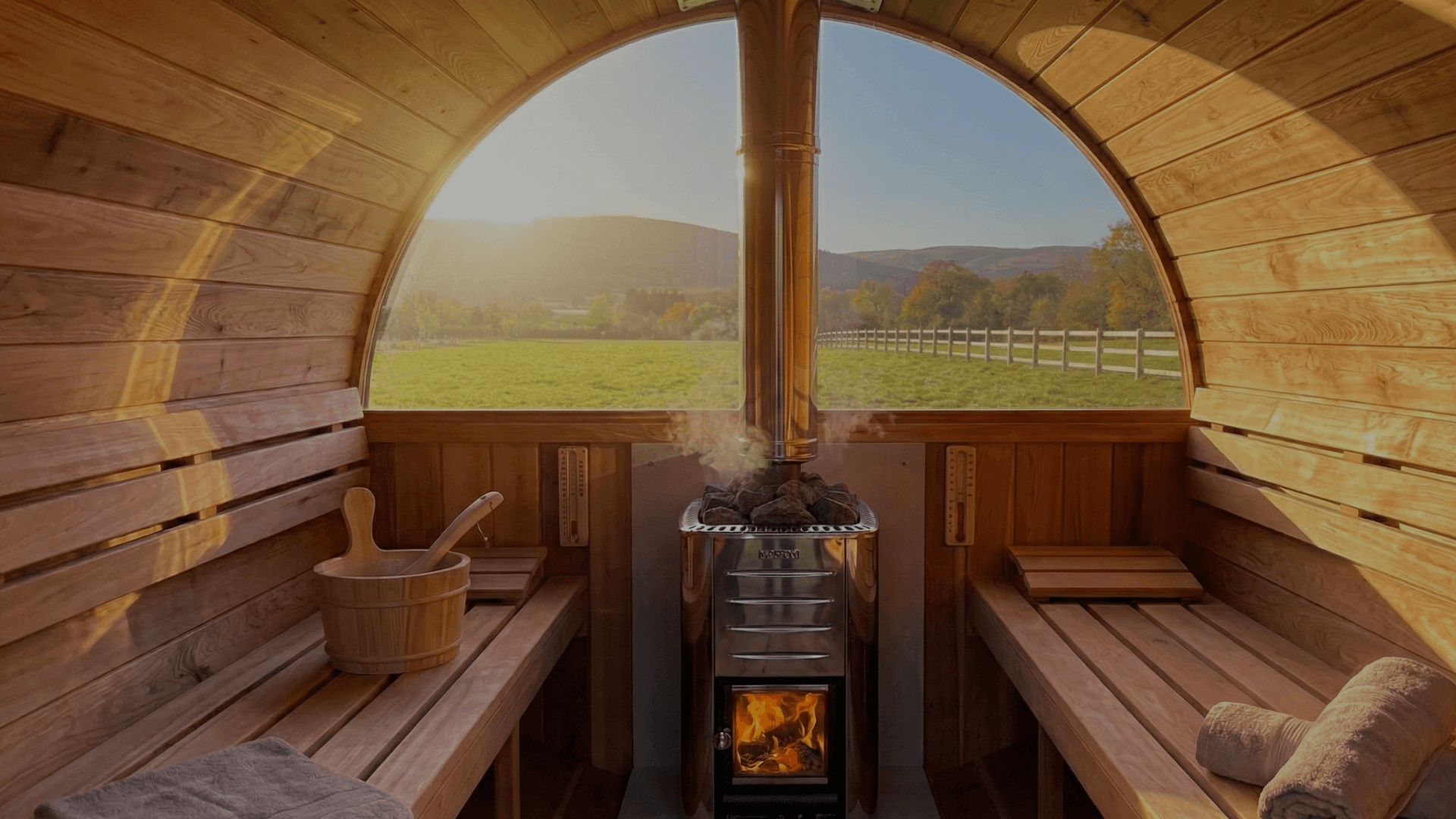 Inside a wooden sauna barrel, a stove with rocks and fire faces a large arched window overlooking a green valley.