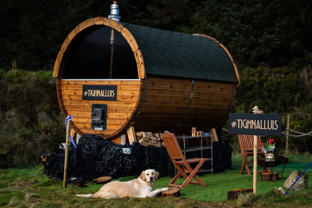 A wooden barrel sauna sits outdoors in a grassy area with a dog lying in the foreground.
