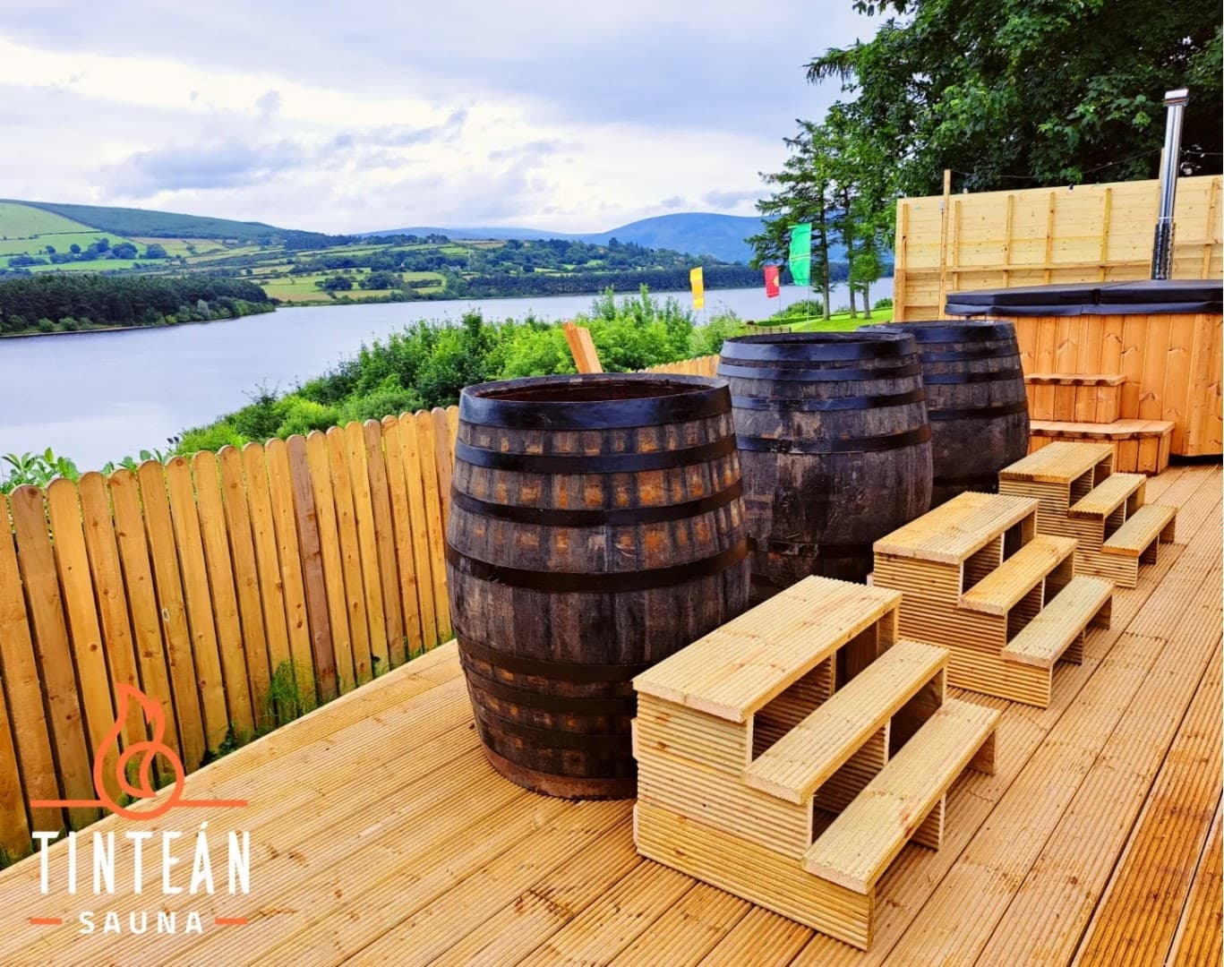 Three large wooden barrels and tiered steps stand on a wooden deck overlooking a lake and hills.