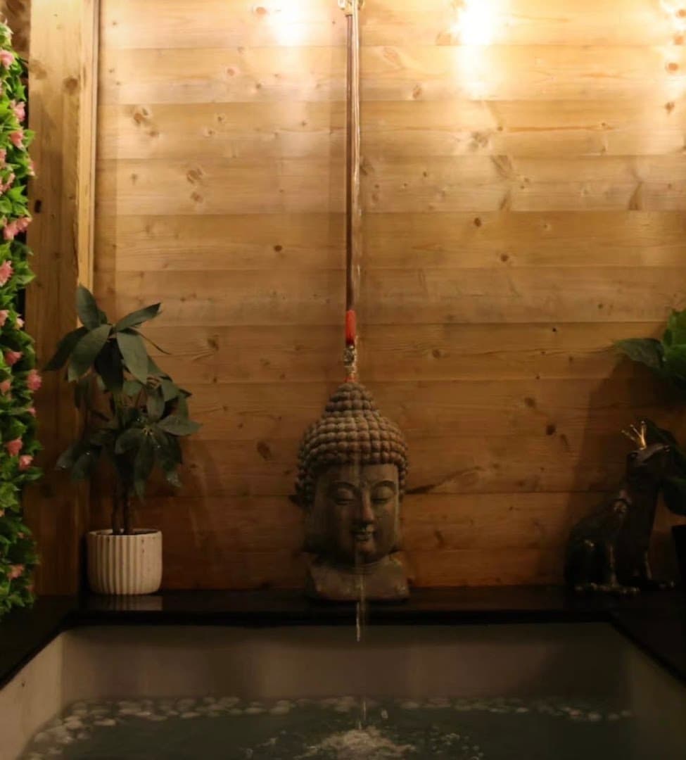 A water fountain with a Buddha statue stands against a wooden paneled wall above a pool.