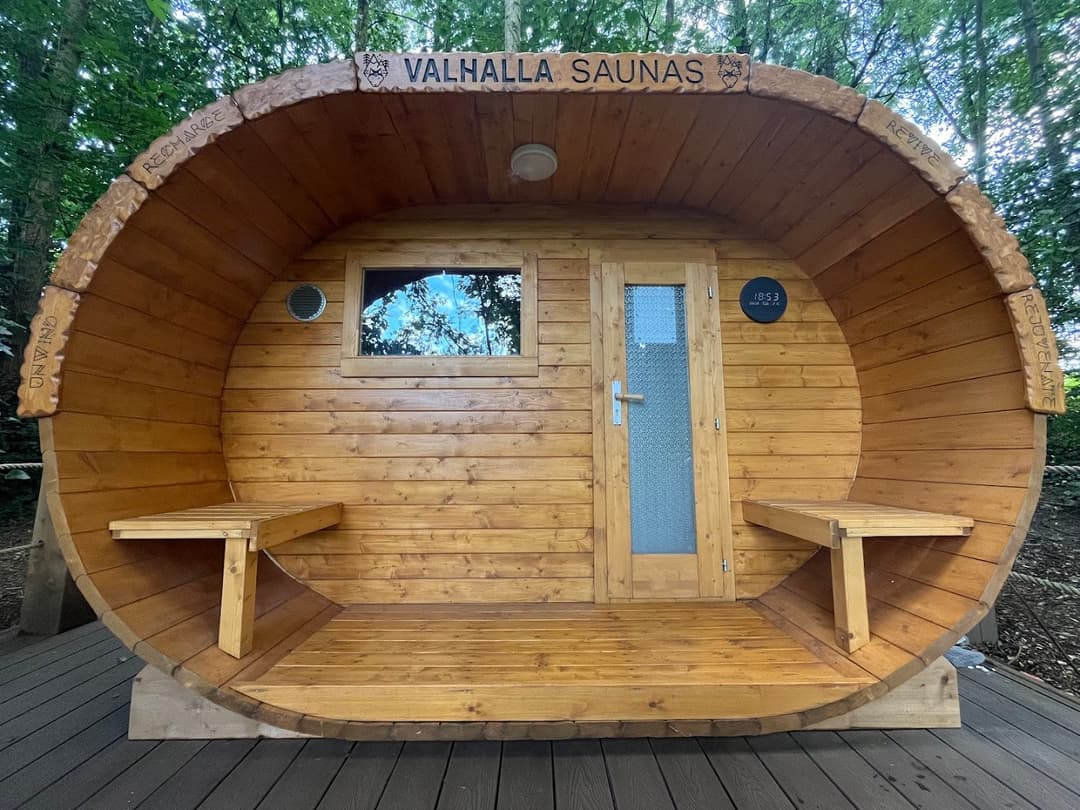 The wooden Valhalla Saunas barrel structure sits on a deck surrounded by trees.