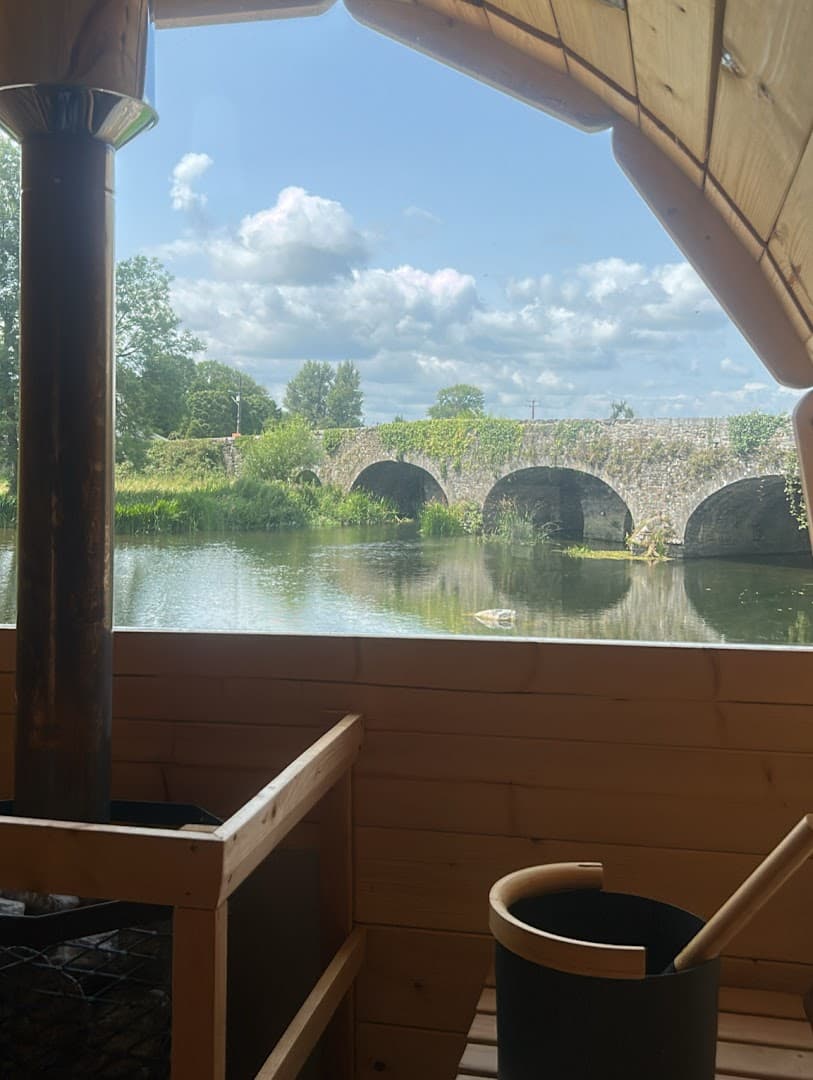 Wooden interior of a sauna overlooking a calm river, green trees, and an old stone bridge.