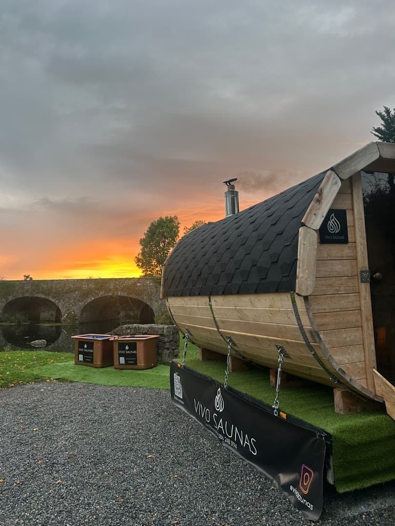 A wooden barrel-style sauna sits outdoors near a stone bridge at sunset.