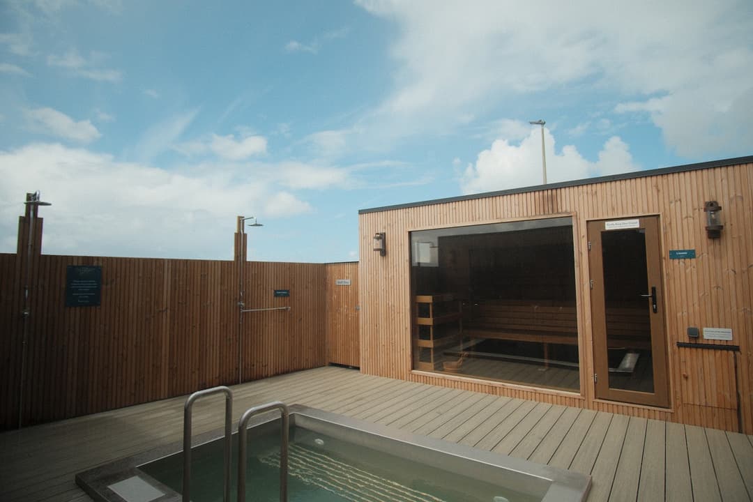 A wooden sauna building with a glass window and a hot tub on a deck, with showers and a wooden fence, under a partly cloudy sky.