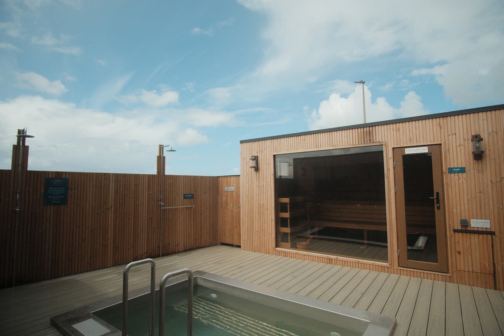 A wooden sauna building with a glass window and a hot tub on a deck, with showers and a wooden fence, under a partly cloudy sky.