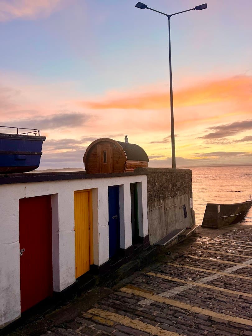 A barrel sauna stands on a pier by the sea during a colorful sunset in Ireland.
