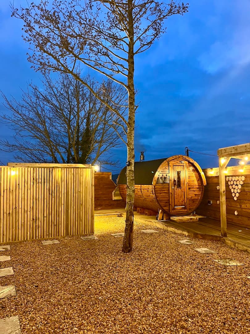 An outdoor barrel sauna on a wooden deck in a gravel courtyard with bare trees at dusk.