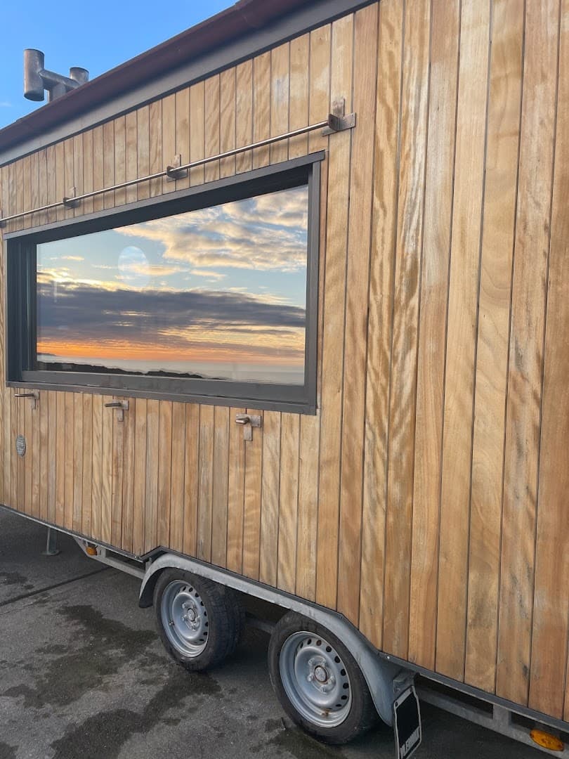 Wooden sauna trailer exterior parked outside with a large window reflecting a sunrise or sunset over the sea.