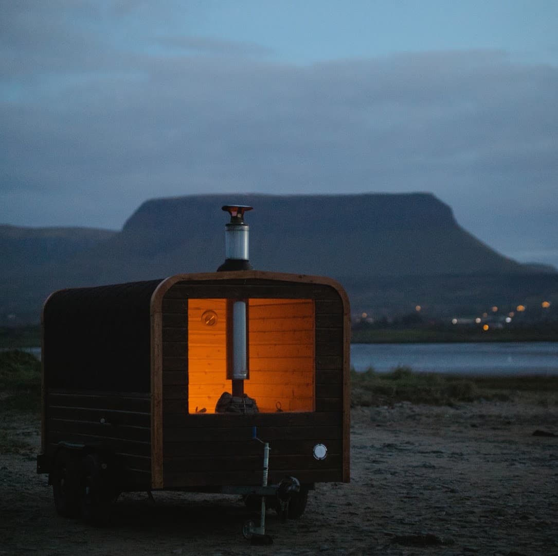 A mobile wooden sauna with an illuminated interior sits on a gravel shore near water, beneath a mountain at dusk.