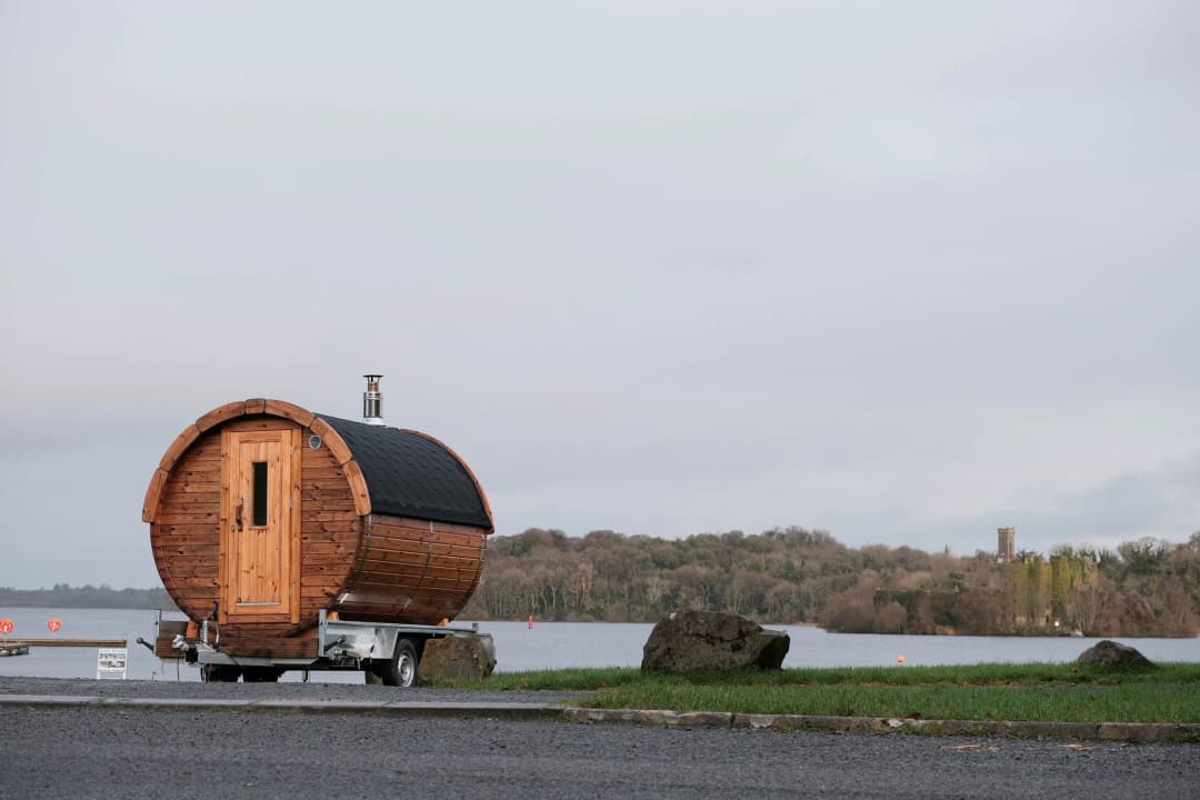A wooden barrel sauna on a trailer sits by a body of water under a cloudy sky.