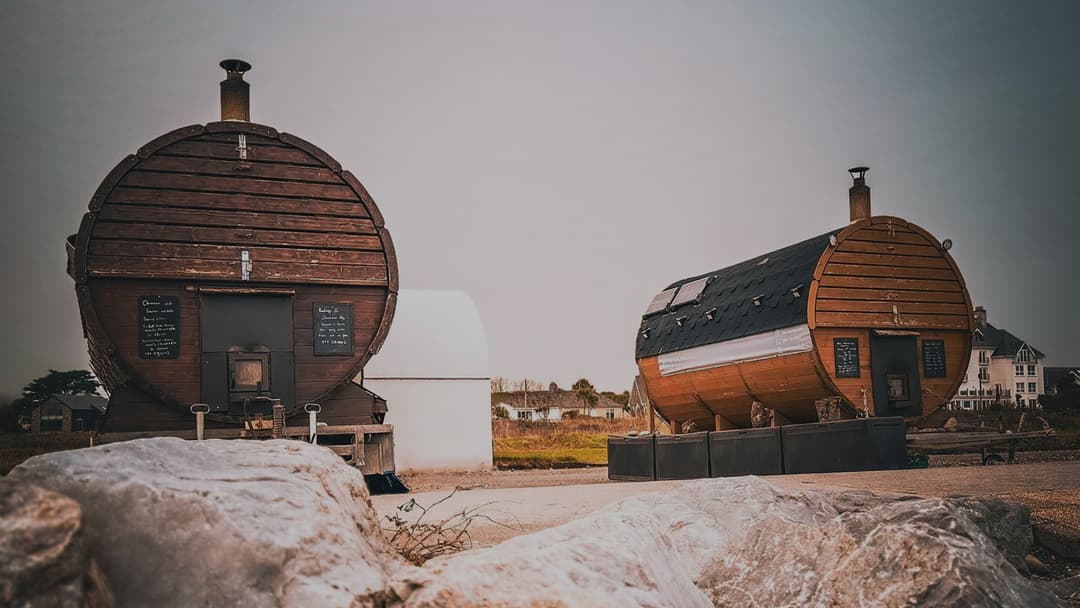 Two barrel-shaped saunas sit outdoors near the coast under a grey sky.