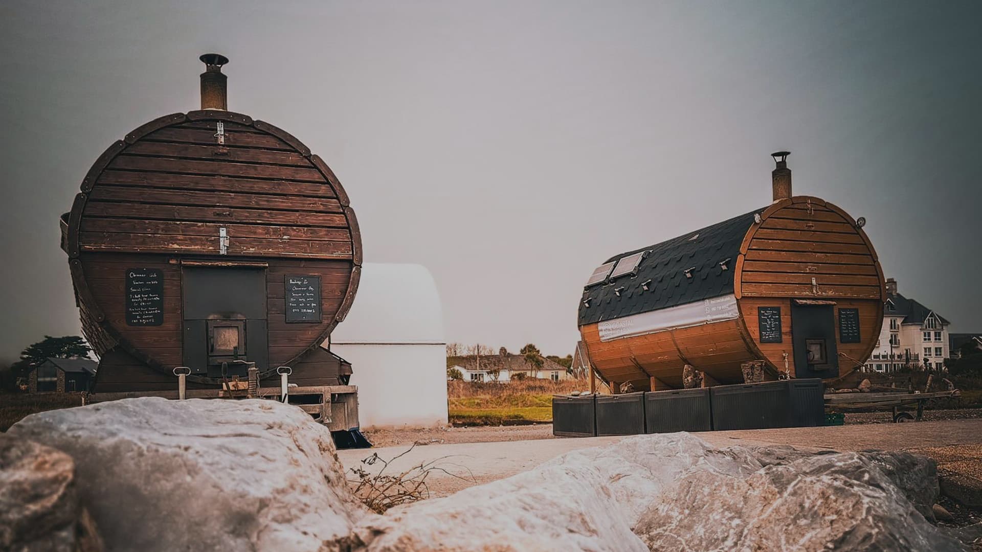 Two barrel-shaped saunas sit outdoors near the coast under a grey sky.