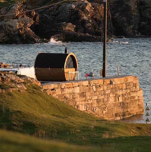 A barrel-shaped wooden sauna sits on a stone pier next to the sea with rocky cliffs in the background.