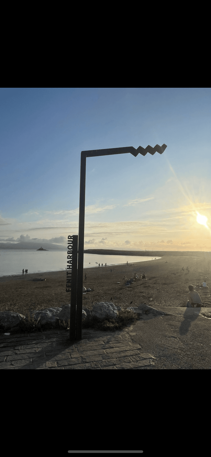 A metal structure stands on a beach near the sea during sunset, with people scattered along the shore.