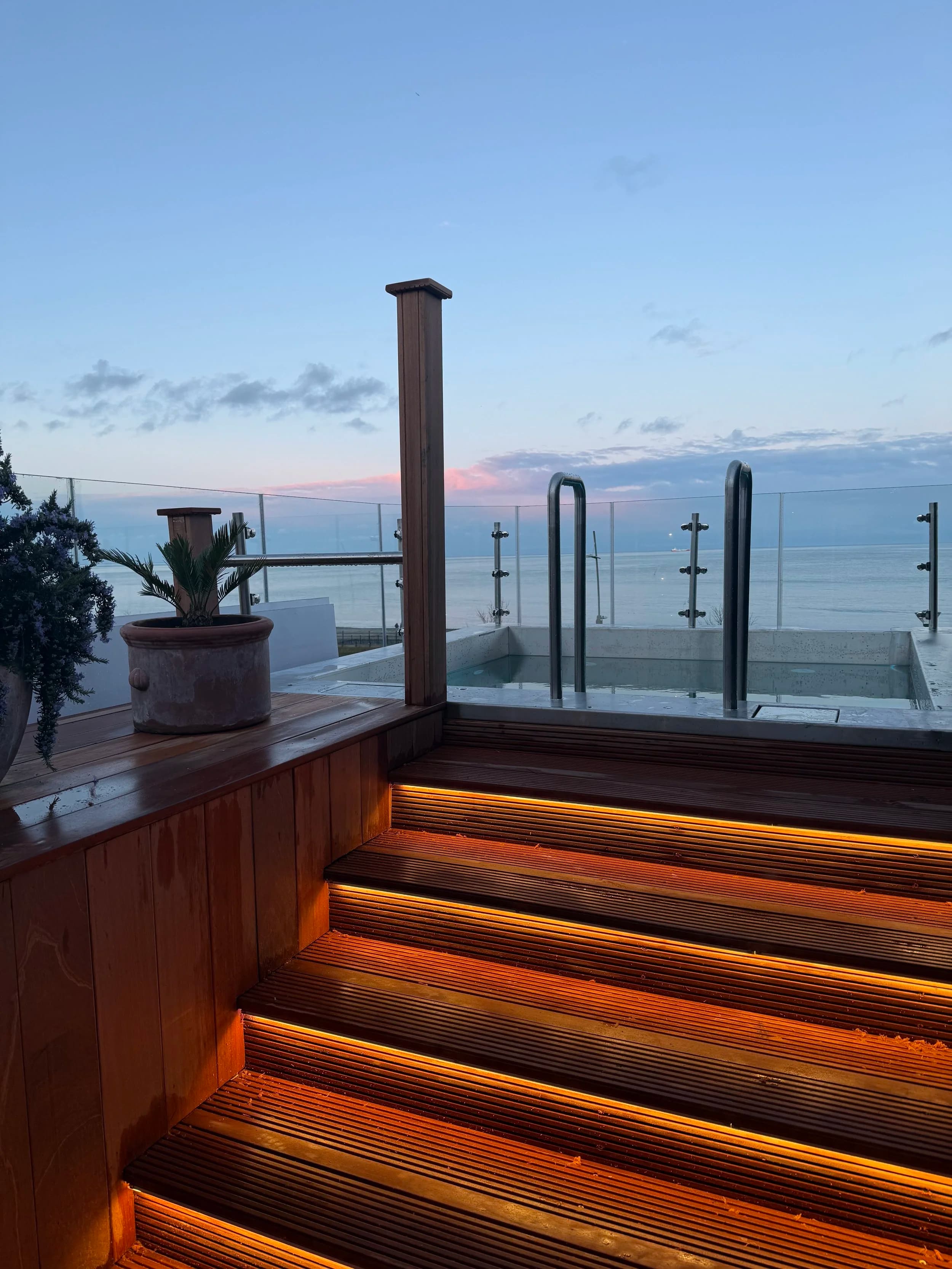 Wooden stairs with amber lights lead up to a hot tub on a rooftop overlooking the sea.