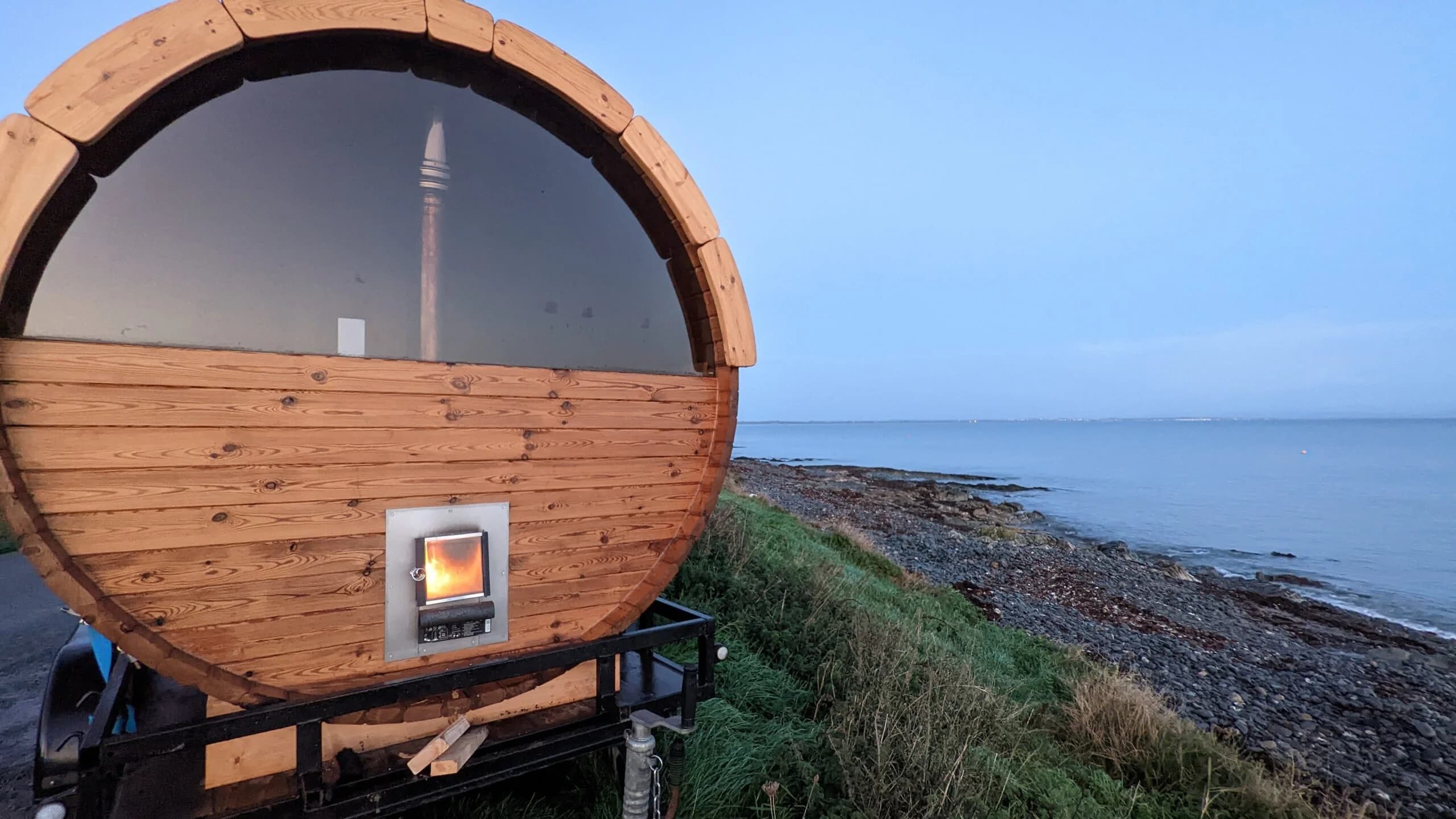 A wooden barrel sauna on a trailer sits by the sea at dusk, with a fire glowing inside the stove.